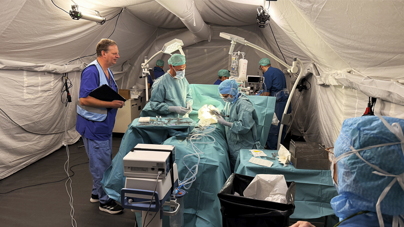 En tillfällig operationssal. Medarbetare helt klädda i blått utför en operation på en fiktiv person. En annan person övervakar operationen.