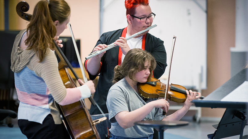 Bild som visar tre ungdomar spelandes instrument, cello, fjol och tvärflöjt.