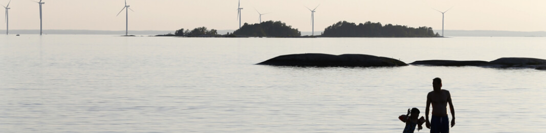 Bild som visar en siluett av en vuxen person och ett barn badandes vid strandkanten i solnedgång med vindkraftverk i bakgrund.