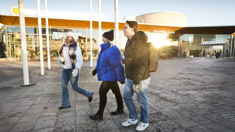 Två kvinnor och en man i vinterjackor går med Karlstads universitets entré i bakgrunden.