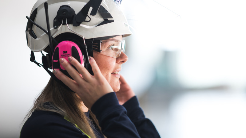 Profil av kvinna med vit hjälm, skyddsglasögon och rosa hörselkåpor som håller händerna på hörselkåporna.
