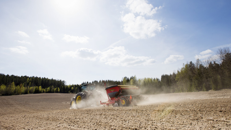 Traktor med såmaskin kör på åker med ett dammoln efter en solig dag.