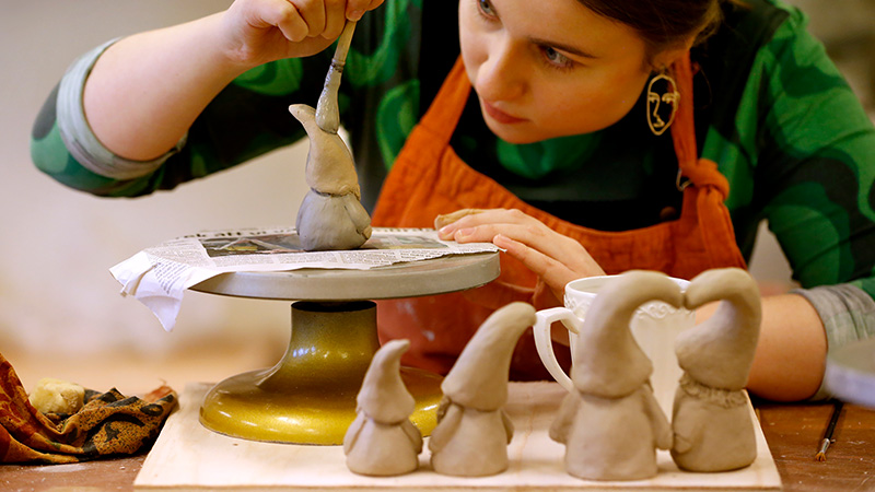 Närbild på kvinna som penslar på obränd lerfigur med stor tomteluva.