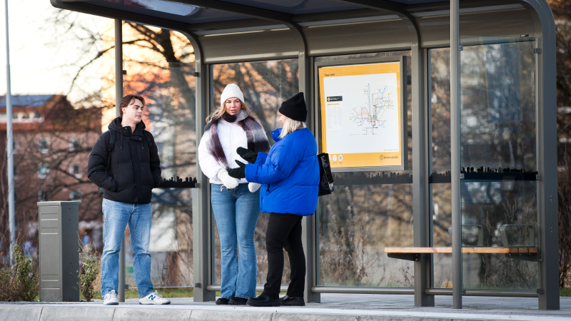 Tre studenter står och pratar medan de väntar på bussen. Hållplats Haga entre och det är kallt ute.