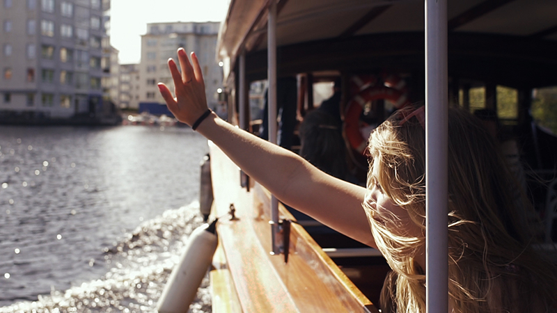 Närbild på kvinna som sitter ombord på båtbuss. Solen skiner och vinden blåser i håret. Hon vinkar.