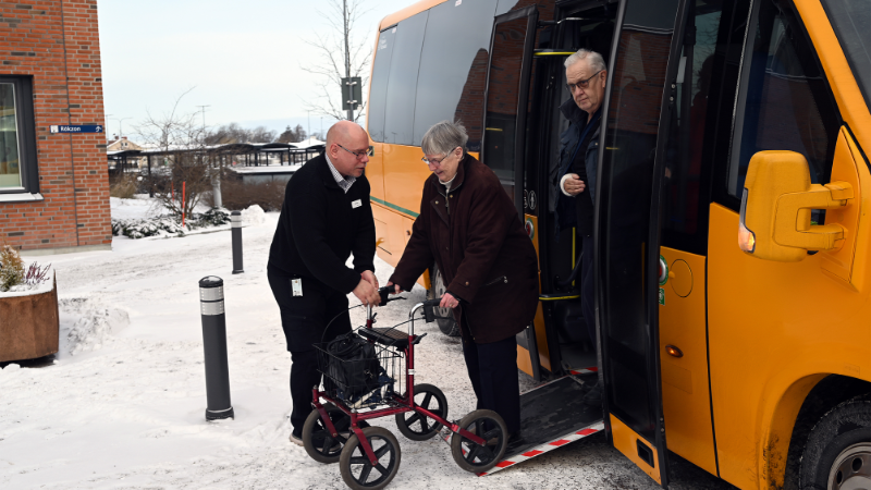 Gul servicelinjebuss parkerad. Föraren hjälper en man och en kvinna av bussen.