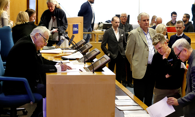 Många personer i föreläsningsal. De vandrar runt och diskuterar samt läser i papper som ligger i högar.