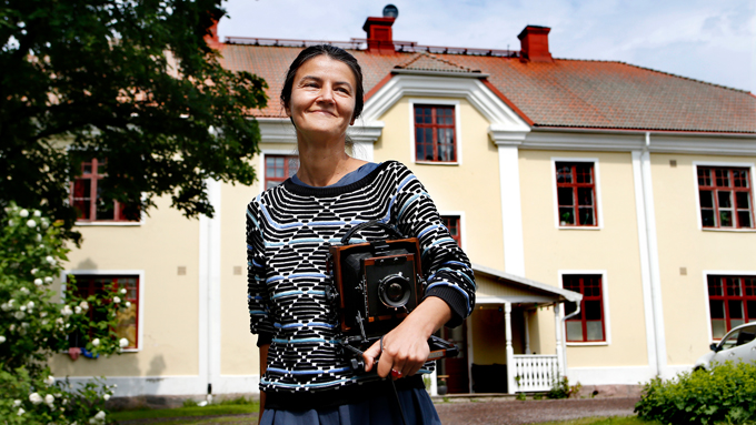 Foto av konstnären Mila Teshaieva framför Gunnerudsgården. Mila ler och håller i en analog kamera.
