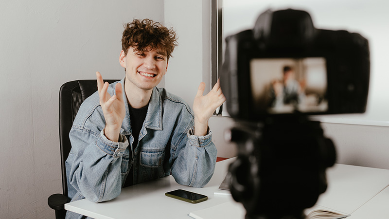 En person sitter vid ett bord och har en kamera uppställd framför sig. 