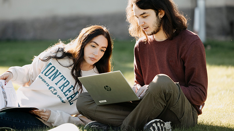 Två personer sitter utomhus och tittar på en laptop som ligger i den ena personens knä. 