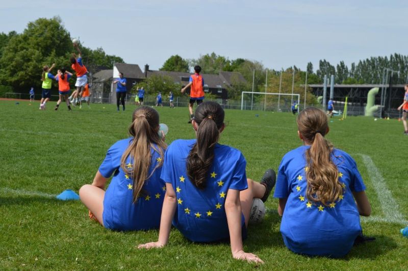 Tre tjejer sitter i gräset vid kanten av en fotbollsplan med EU loggan på sin tröja.