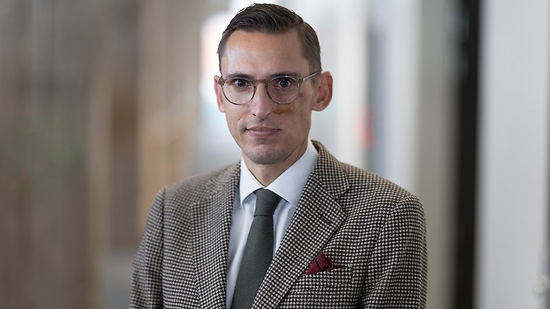 En man med kostym och glasögon tittar in i kameran.