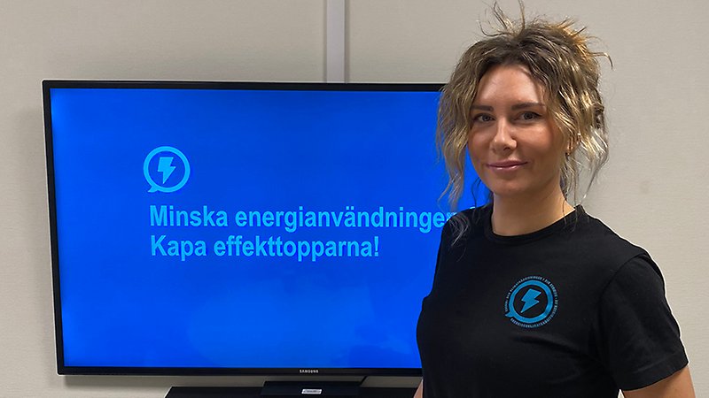 Kvinna med uppsatt här står vid en TV-skärm och tittar in i kameran.