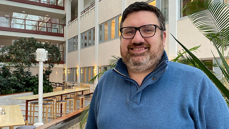 En man med skägg och glasögon tittar in i kameran och ler.